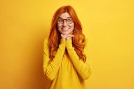 Joyful surprised redhead young woman keeps hands under chin shows white teeth feels glad listens with great interest interlocutor dressed in casual jumper isolated over vivid yellow background